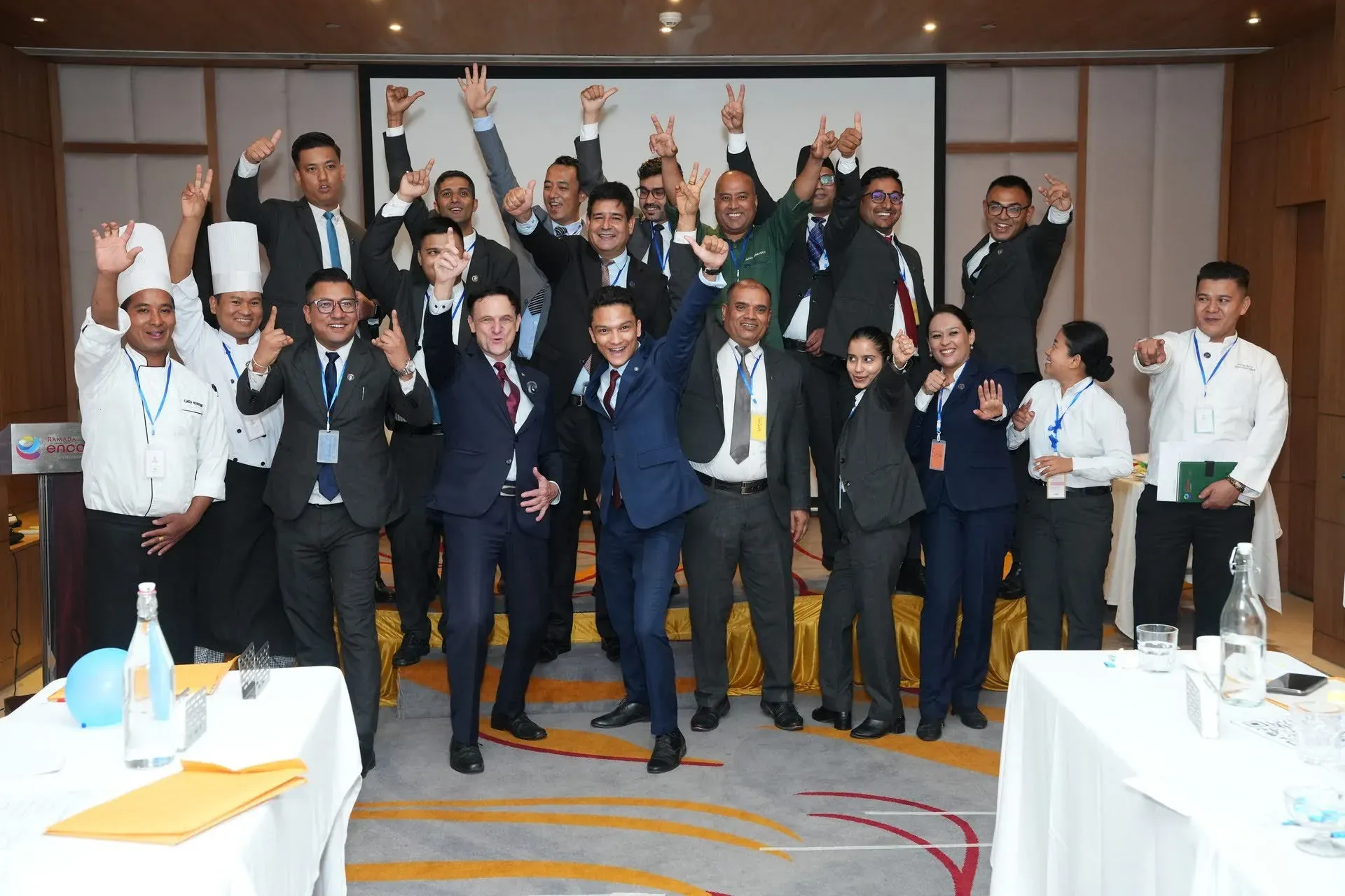 The full Ramada Encore cohort, hotel staff in uniform and in suits, hands raised at the close of the programme.