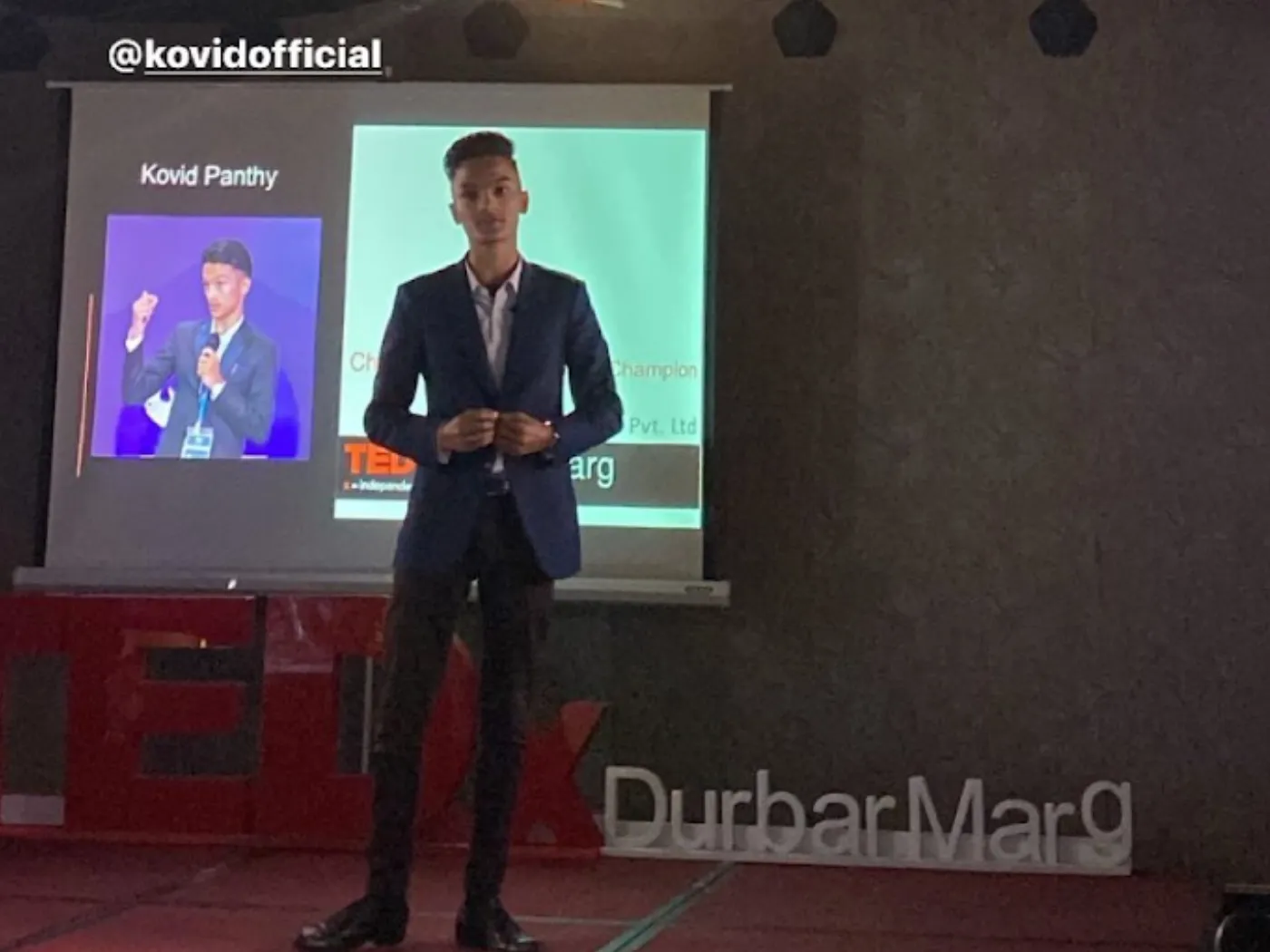 Kovid Panthy speaking on the TEDx Durbarmarg stage, his name and photograph on the screen behind him.