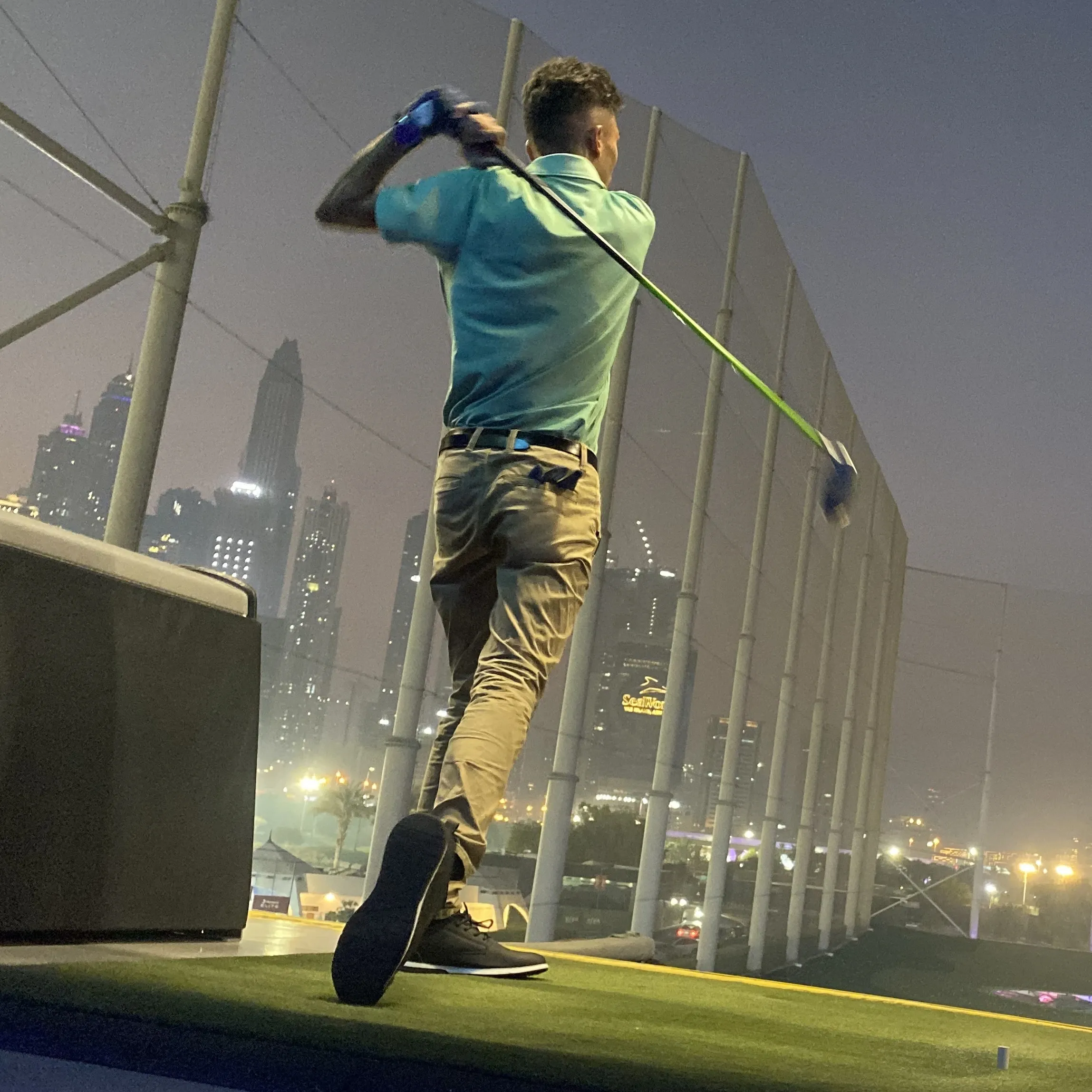 Kovid Panthy at the top of his backswing on a floodlit driving range, a city skyline behind.