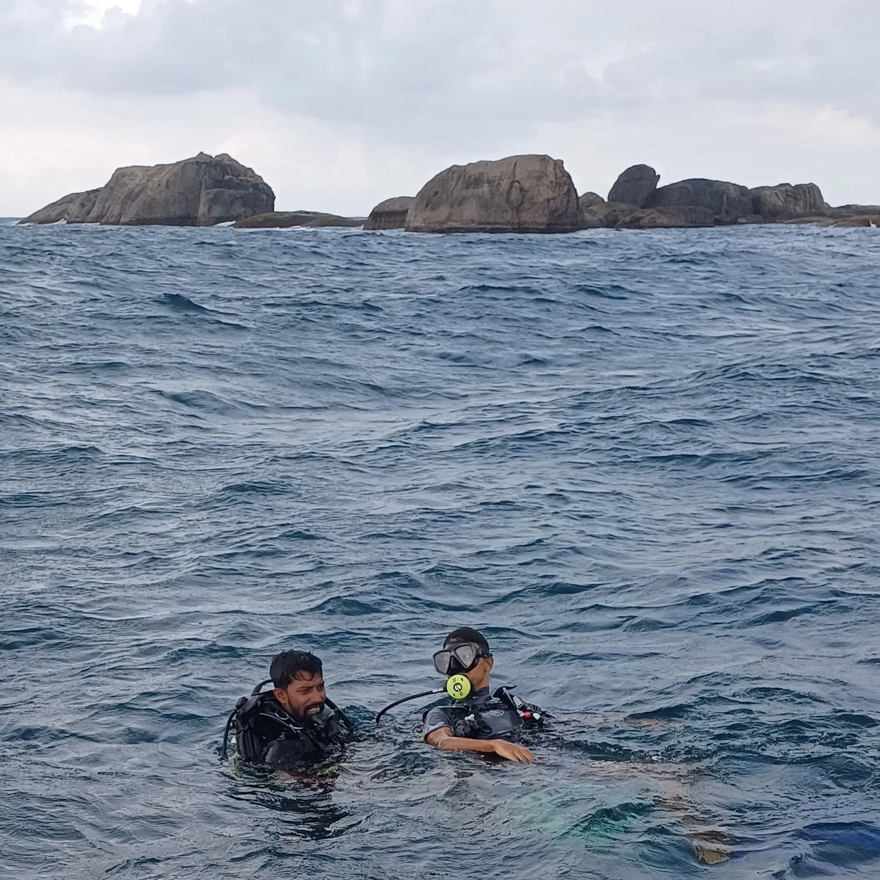 Two divers in the water in full kit, a rocky headland behind them.
