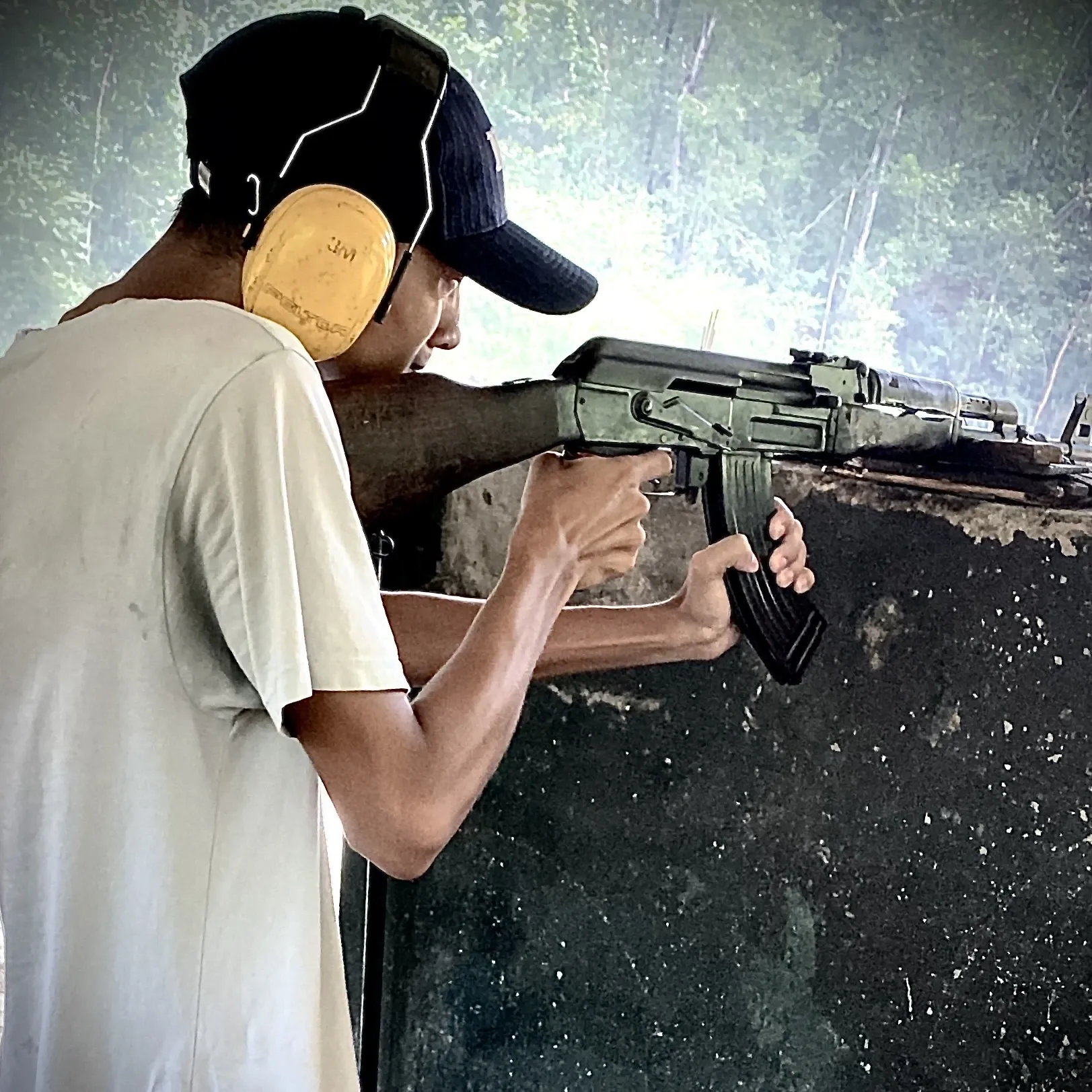 Kovid Panthy at an outdoor shooting range in ear defenders and a cap, rifle shouldered at the bench.