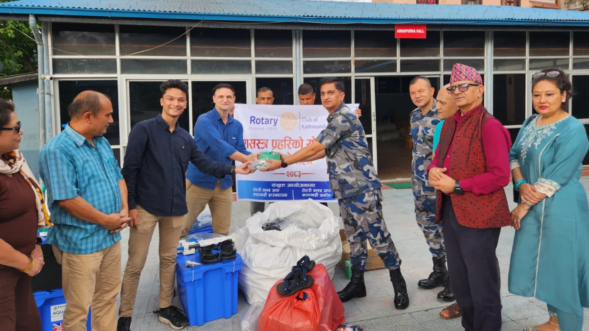 Kovid Panthy handing rescue equipment to an Armed Police Force officer beneath a Rotary Club of Kathmandu banner, relief crates and dry bags stacked in front.