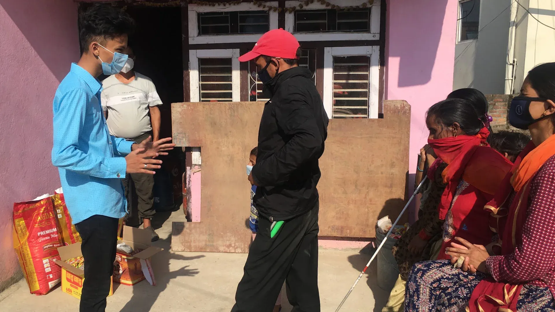 Sacks and boxes of rations being handed out during the Covid-19 pandemic, recipients waiting alongside, one with a white cane.